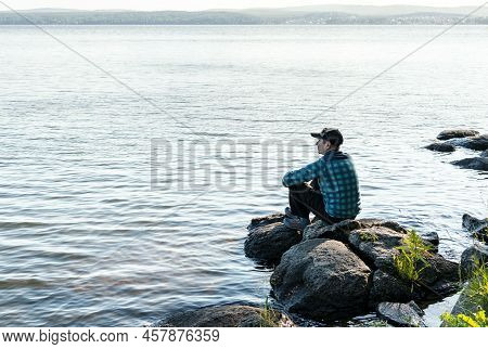 Young Man In A Cap Looking At The View Of The Lake Or River Sitting On The Stones Enjoying Nature La