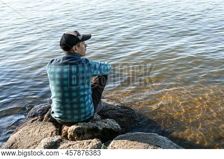 Young Man In A Cap Looking At The View Of The Lake Or River Sitting On The Stones Enjoying Nature La
