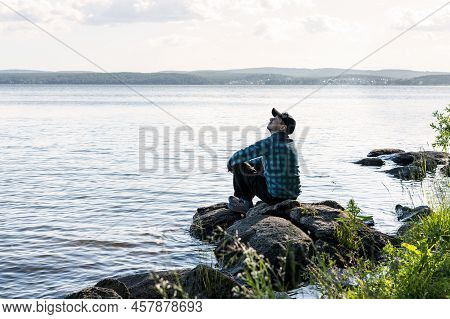 Young Man In A Cap Looking Up Sitting On The Stones On Shore Of Lake Or River Enjoying Nature Landsc