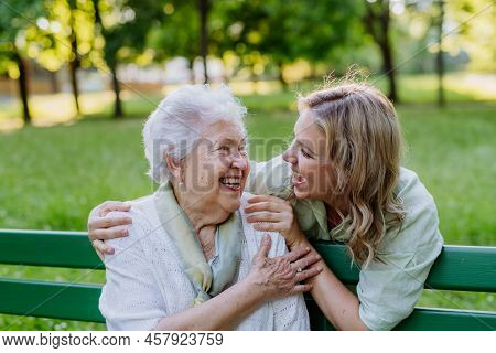Adult Granddaguhter Helping Her Grandmother To Use Cellphone When Sitting On Bench In Park In Summer