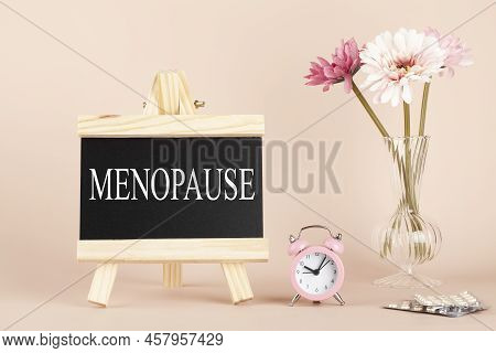 Menopause Word Written On Black Board, Flowers, Clock And Pills On Beige Background. Women Health An