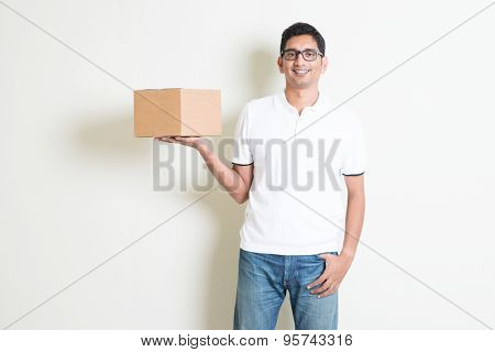 Indian man smiling and holding delivery courier box, standing on plain background with shadow. Asian handsome guy model.