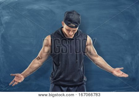 Muscular man in black clothes and cap with his head lowered on blue chalkboard background. Fitness and working out. Healthy living. Sport achievements.