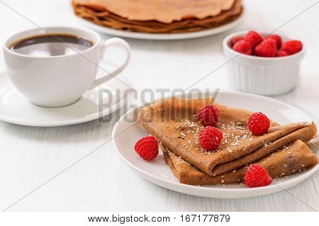 Healthy vegan carob crepes decorated with flaked coconut. Whole wheat chocolate crepes with fresh raspberries on white plate and cup of tea and coffee on white wooden background. Healthy breakfast concepthorizontal