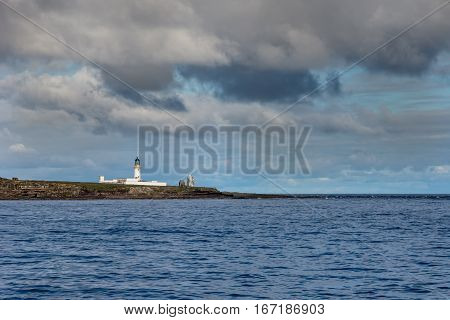 Orkneys Scotland - June 5 2012: The white lighthouse complex on north end of Stroma Island on a rocky shore under heavy cloudy sky. Dark blue sea. Dominant color is blue but by a line of brown rocks.