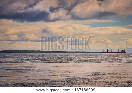 Orkneys Scotland - June 5 2012: Bulk carrier ship plows through the churning waves where the Atlantic meets the North Sea. Narrow green band of Stroma Island between gray sea and cloudy skies with blue patches.