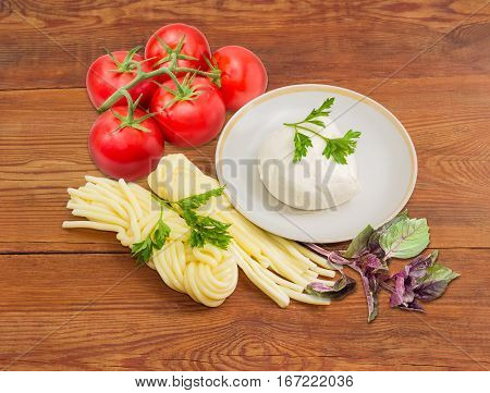 Piece of the fresh mozzarella cheese on the saucer two portion of the mozzarella cheese twisted to form a plait cluster of tomatoes and twigs of parsley and purple basil on an old wooden surface