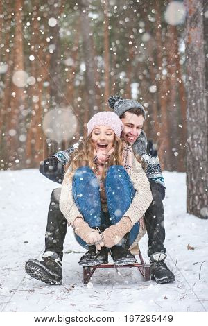 Young couple in winter forest sledding, laughing during snowfall