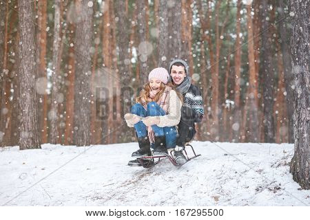 Young couple in winter forest sledding, laughing during snowfall