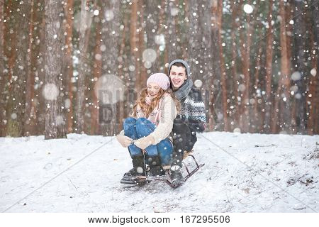 Young couple in winter forest sledding, laughing during snowfall