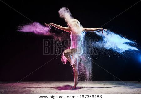 Woman in beige body posing on a tiptoe in cloud of pink and yellow dust streching her arms gracefully
