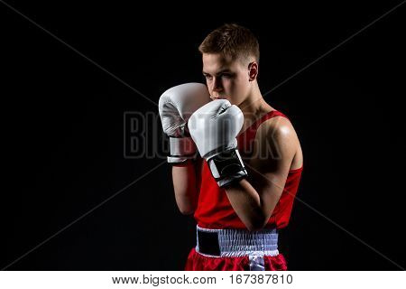 Young handsome boxer sportsman in red boxer suit and white gloves standing on black backgound. Copy space.