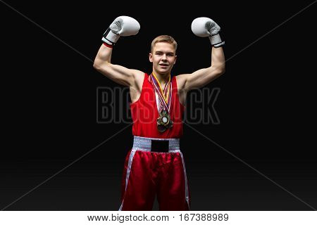 Happy young handsome boxer sportsman in red boxer suit and white gloves with medals standing on black backgound. Winner. Copy space.