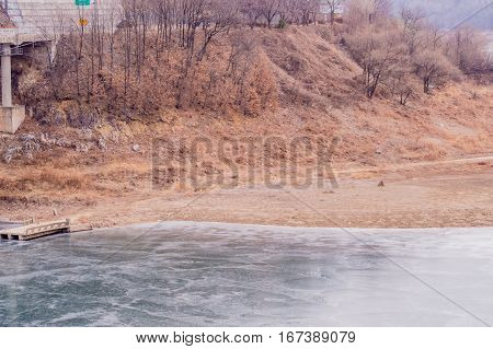 Landscape of a river with a piece of a small concrete bridge extended over the far side of the river and tall brown grass and trees in the background
