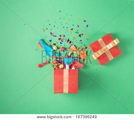 Red gift box with various party confetti balloons streamers noisemakers and decoration on a green background. Colorful celebration background. Flat lay.