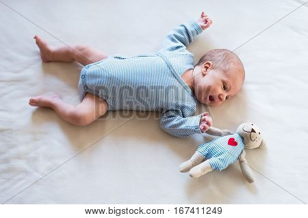 Cute newborn baby boy in blue striped onesie lying on bed, teddy bear toy with red heart next to him