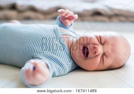 Cute newborn baby boy in blue striped onesie lying on bed, crying. Close up.