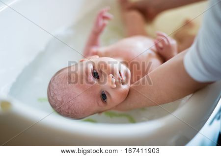 Unrecognizable mother holding her baby son, bathing him in small white plastic bath. Close up.