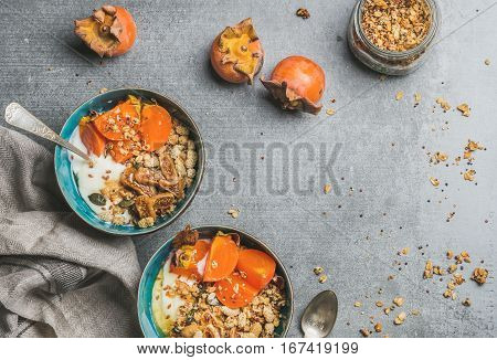 Healthy vegetarian breakfast. Oatmeal, quinoa granola with yogurt, dried fruit, seeds, honey, fresh persimmon in bowls over grey background, top view, copy space. Gluten free, allergy-friendly concept