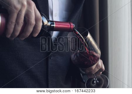 Horizontal close up of Caucasian man in black suit and white shirt pouring red wine into a tall glass in a bar