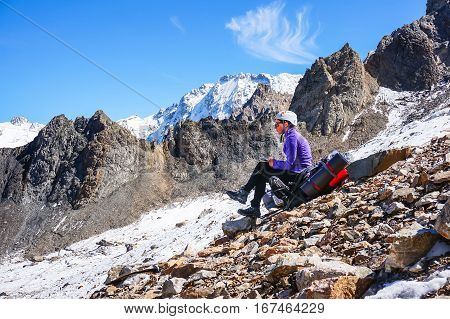 Young female mountaineer in a helmet resting sitting on a backpack and writes the passed route. Against the background of a chain of high rocky mountains and glacier