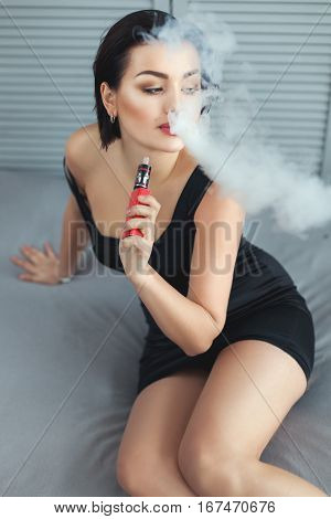 Erotic woman sitting up in bed smoking and produces smoke in her hand electronic cigarette.