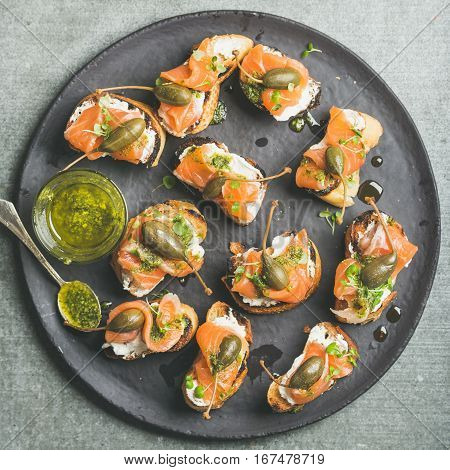 Homemade salmon crostini with cream-cheese, watercress, capers and pesto suace in round black slate stone plate over grey background, top view, square crop