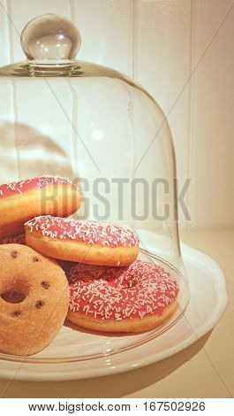 Clopseup assorted donuts on a plate covered glass bell. White painted wooden planks on background. Warm light