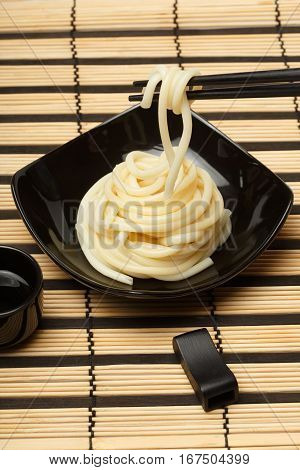 Spaghetti macaroni taking from black ceramic dish by chopsticks at striped bamboo mat background with sake cup and chopsticks rest near.