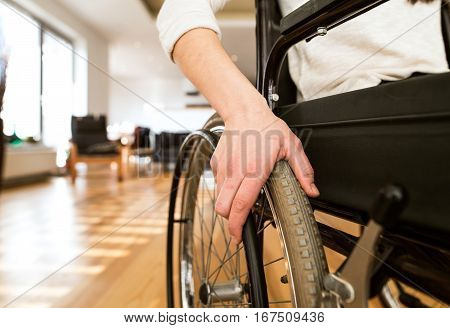 Unrecognizable young disabled woman in wheelchair at the at home in her living room. Close up of her arm laid on wheel.
