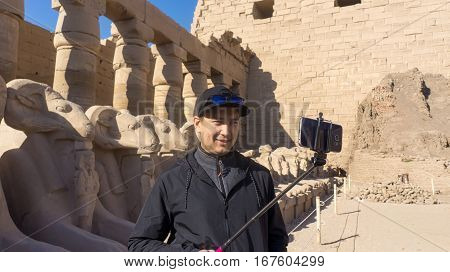Happy man on holidays visiting ancient Egyptian temple in Luxor Egypt taking selfie shot using his mobile phone and selfie stick