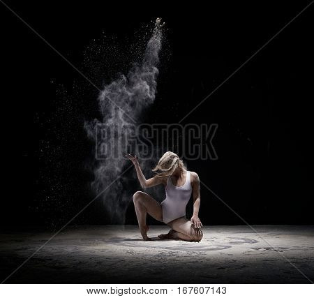 Female sexy long haired blonde in beige bodysuit sitting gracefully on black background white dust all over body studio shot