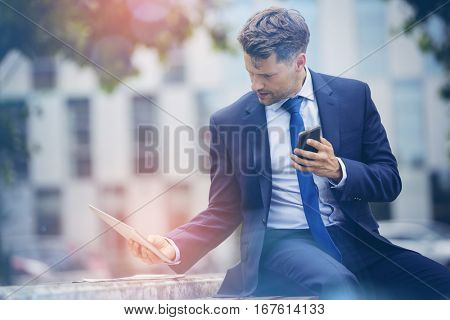 Businessman holding mobile phone while using digital tablet on retaining wall
