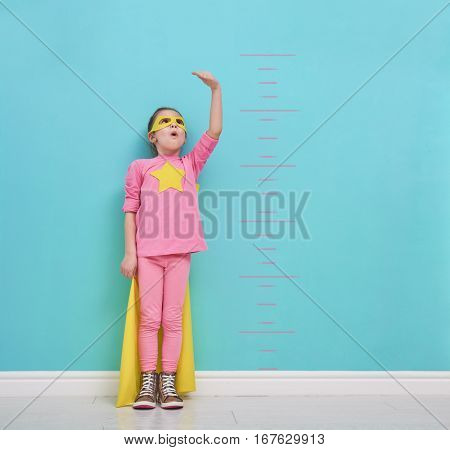 Little child plays superhero. Kid measures the growth on the background of bright blue wall. Girl power concept. Yellow, pink and  turquoise colors.