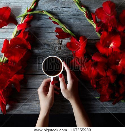 Cup of hot black coffee in female hands on a wooden vintage desk with summer red flowers gladioli. The concept of coffee on a summer day.