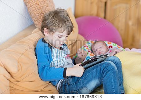 Preschool kid boy playing games tablet computer during babysitting. Cute newborn baby looking on brother. Adorable kids and children at home.
