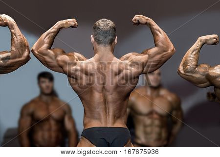 Bodybuilder flexing back and biceps. Muscular athlete posing on stage. Top sportsmen at bodybuilding competition. Opponents are watching.