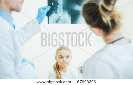 Little Girl Awaiting Medical Diagnosis in Front of Two Hospital Physicians with X Ray Image.