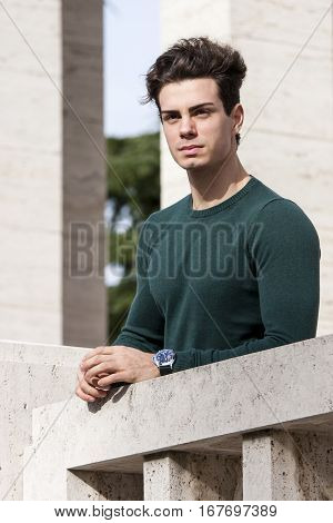 Stylish hair young man outdoors on the ledge, tight knit. A beautiful young European (Italian) man is outdoors resting on the ledge. The boy wears a tight knit long-sleeved green.