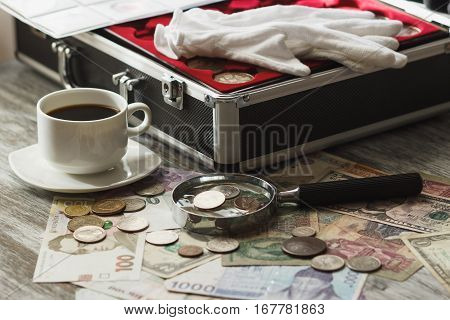 Different collector's coins and banknotes with a magnifying glass box for coins and cup of coffee soft focus background