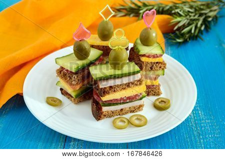 Mini sandwiches (canape) with fresh cucumbers ham cheese olives salami black bread on a white plate on blue background.
