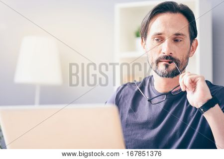Let me think. Pleasant concentrated adult man holding his glasses and using laptop while working at the table