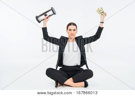 Young businesswoman sitting on floor and holding hourglass and money on white