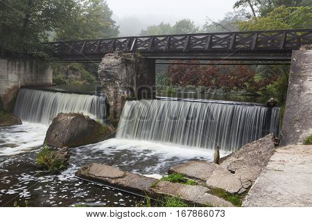 Wooden bridge across the river with waterfall. Foggy morning.