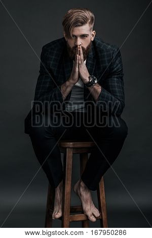 Confidence and charisma. Full length of attractive young man keeping hands clasped and looking at camera while sitting against grey background