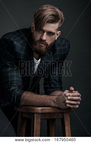 e looking in your heart. Confident man in casual clothing looking at camera while leaning on stool against grey background