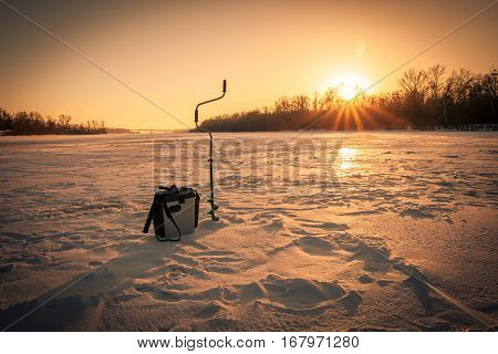 Ice fishing scene with box drill and a rod on river during sunset