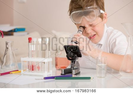 Cute elementary schoolboy looking into microscope at his desk at home. Young scientist making experiments in his home laboratory. Indoors. Child and science. Education concept.