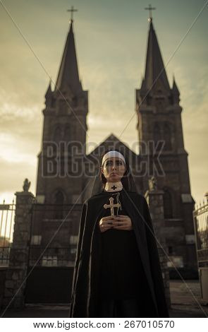 Beautiful Young Nun In Black Cassock And Cloak Is Standing With Cross In Her Hands On Temple Backgro