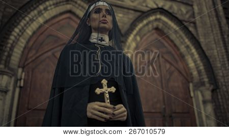 Beautiful Young Nun In Black Cassock, Cloak And Transparent Cape Is Standing And Praying With Cross 